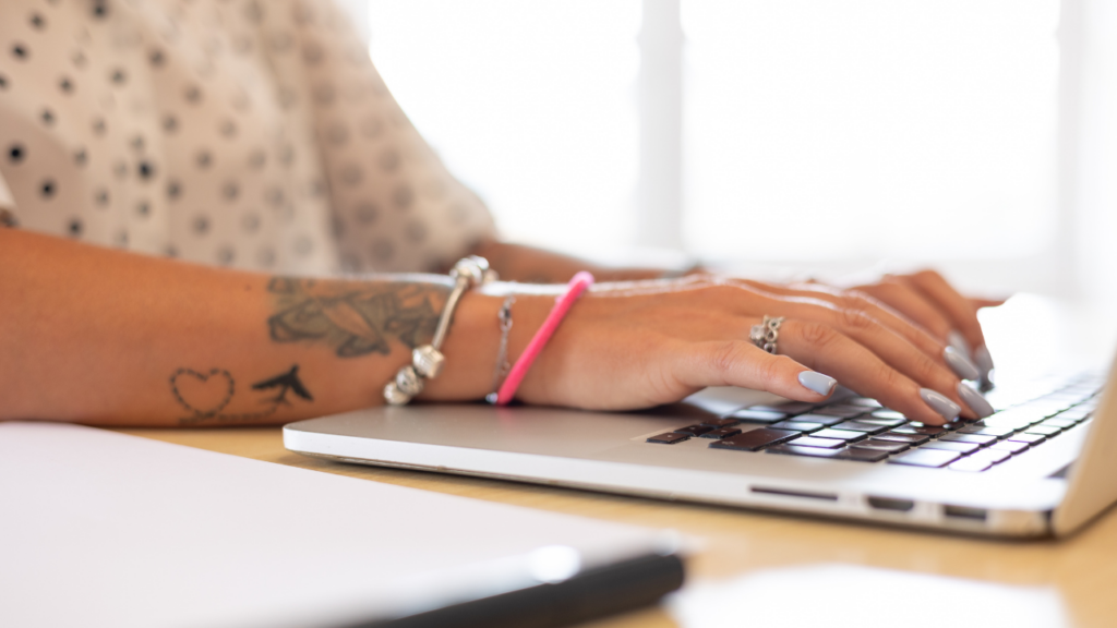 Mani di una persona con tatuaggi e braccialetti che digitano sulla tastiera di un laptop, su una scrivania con luce naturale sullo sfondo. La persona scrvie come scrivere una lettera di presentazione